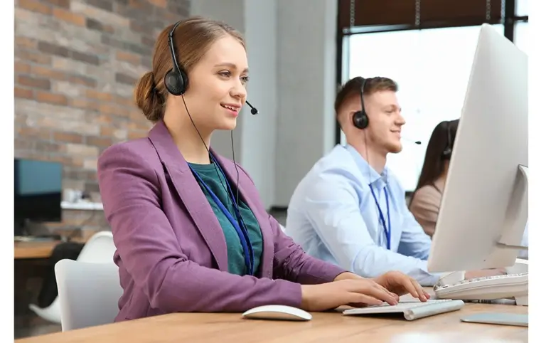 Pracovníčka supportu vo fialovom saku sedí za počítačom a poskytuje telefonickú podporu zákazníkom. V pozadí sedí jej kolega, ktorý poskytuje rovnakú službu.